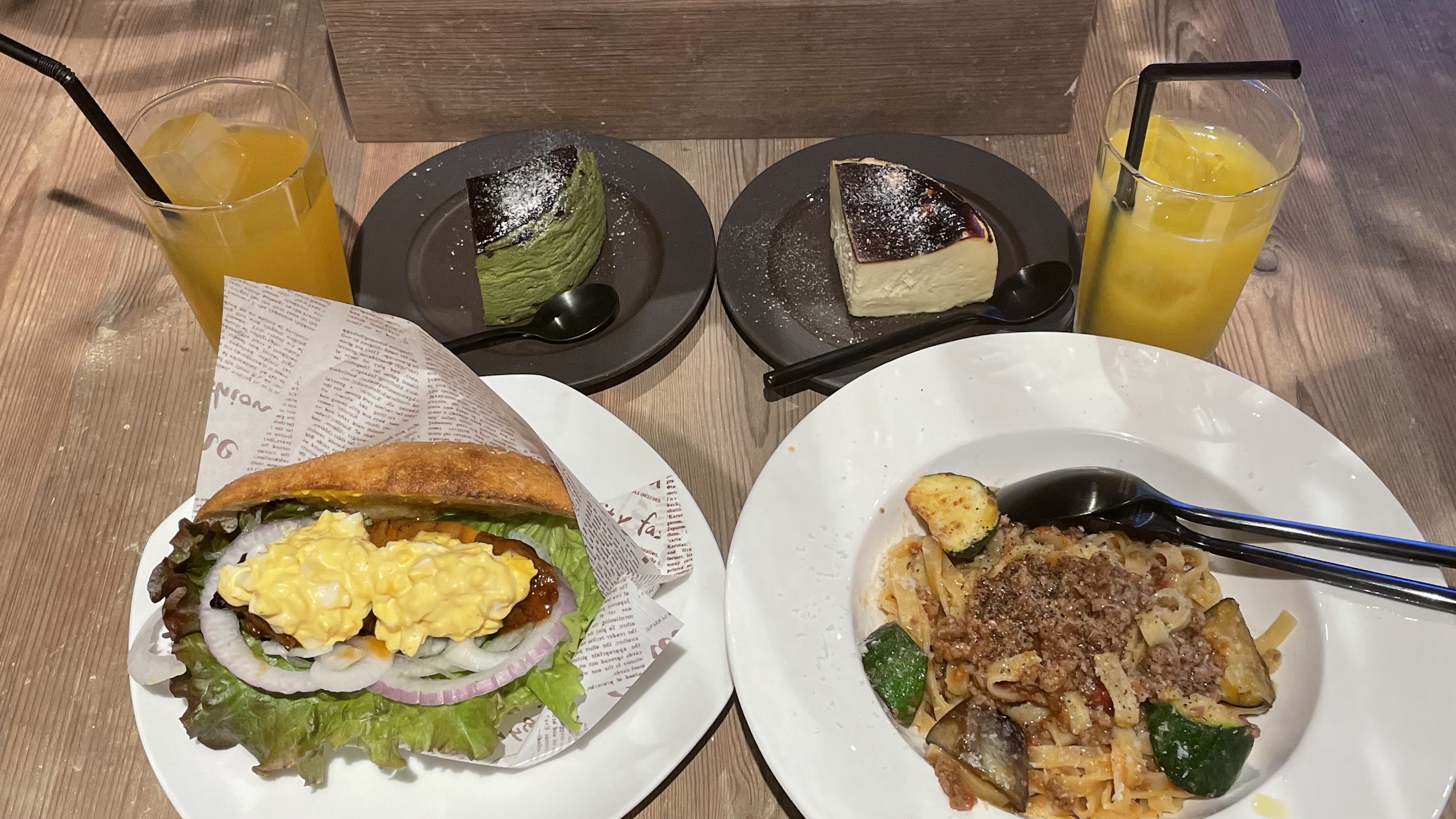 木目テーブルに並ぶパスタ、サンドイッチ、抹茶ティラミス、チーズケーキとドリンク 2 杯